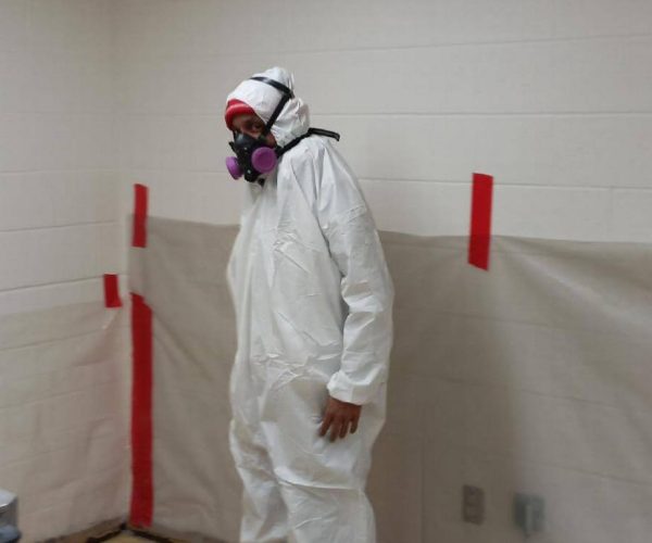 A man in a hazmat suit standing in a room, wearing gloves and a mask, with a caution sign on the wall.
